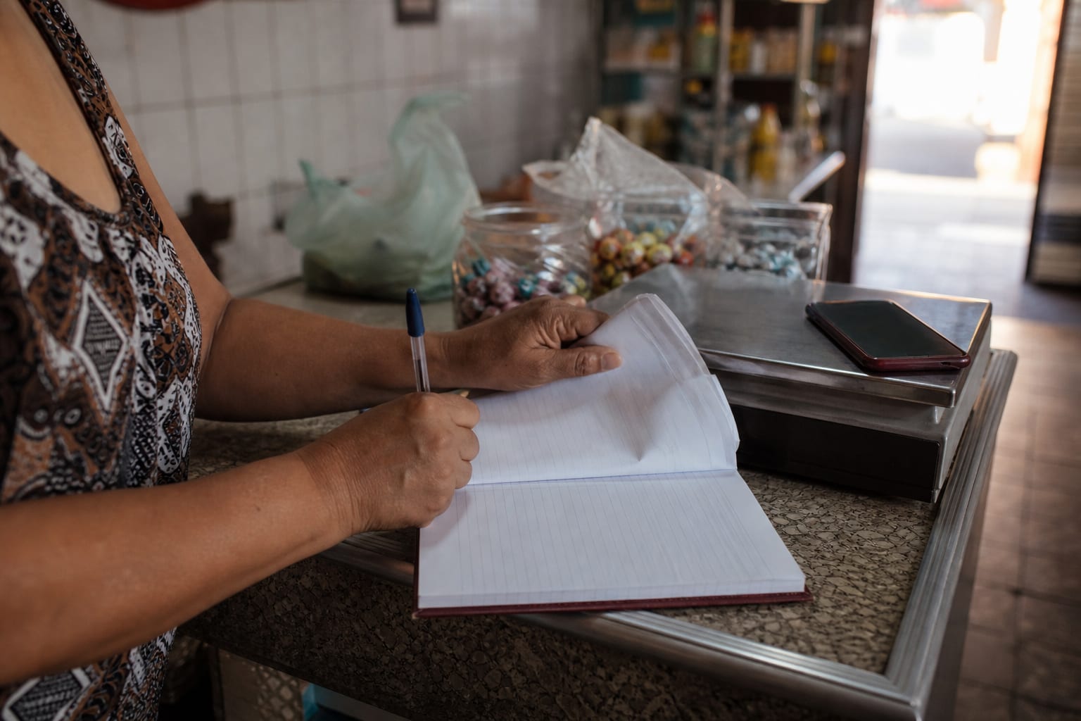 Mãos de comerciante entre um caderno de fiado aberto e um celular no balcão de mercearia