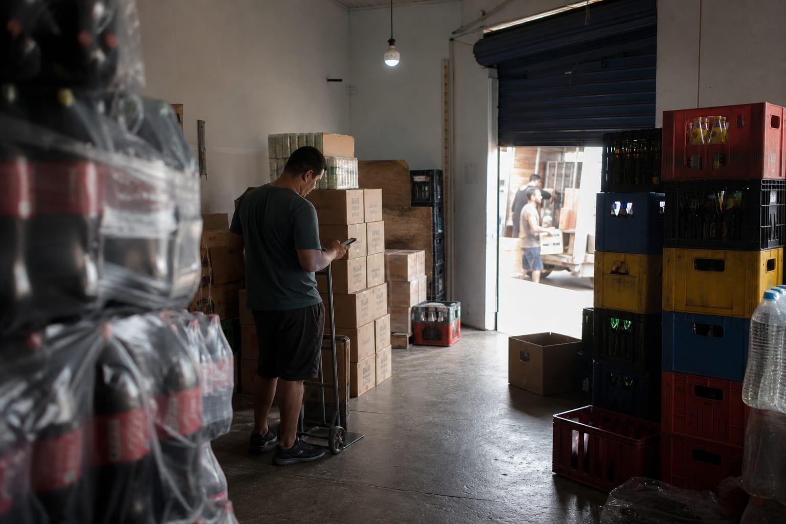 Comerciante confere caixas de bebida no estoque dos fundos com o celular na mão